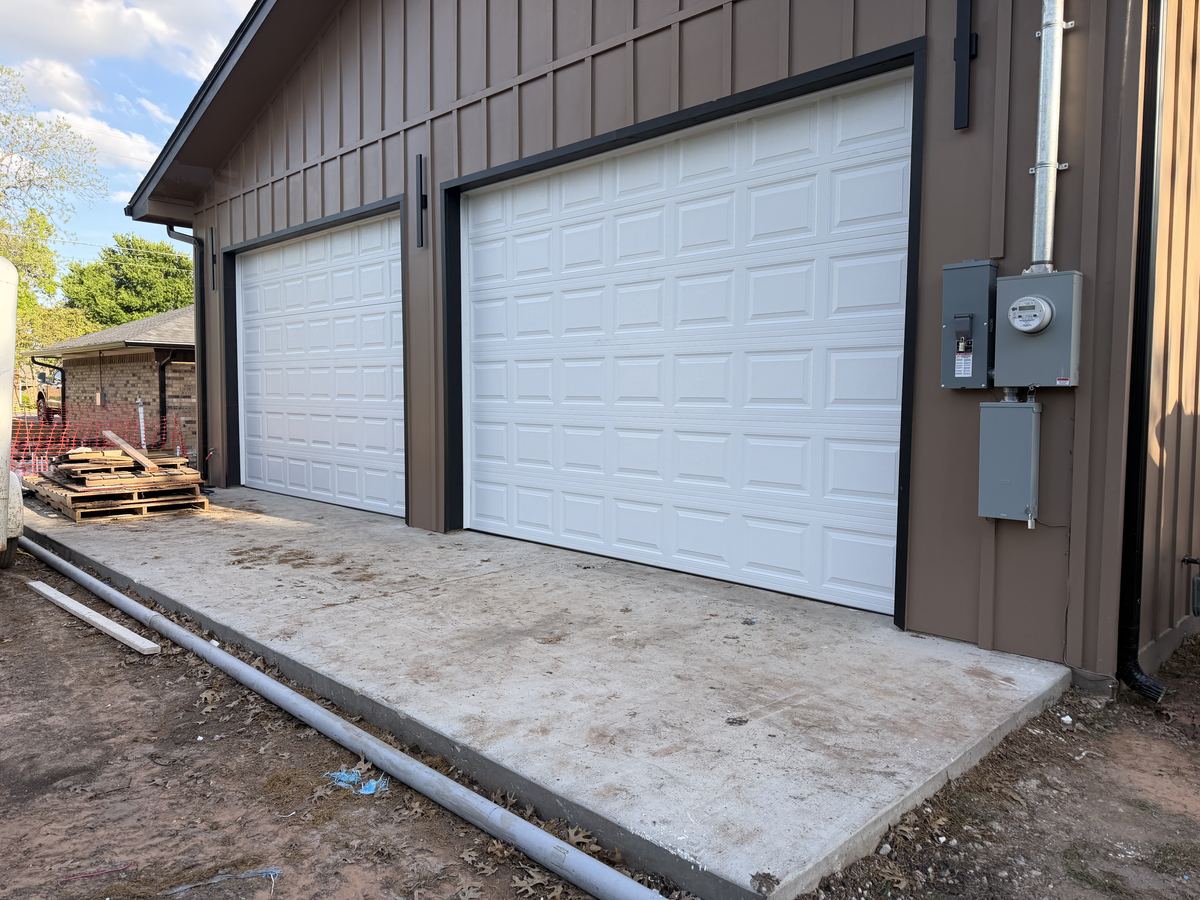 Two white raised-panel overhead doors on a brown board-and-batten shop building with a new concrete apron
