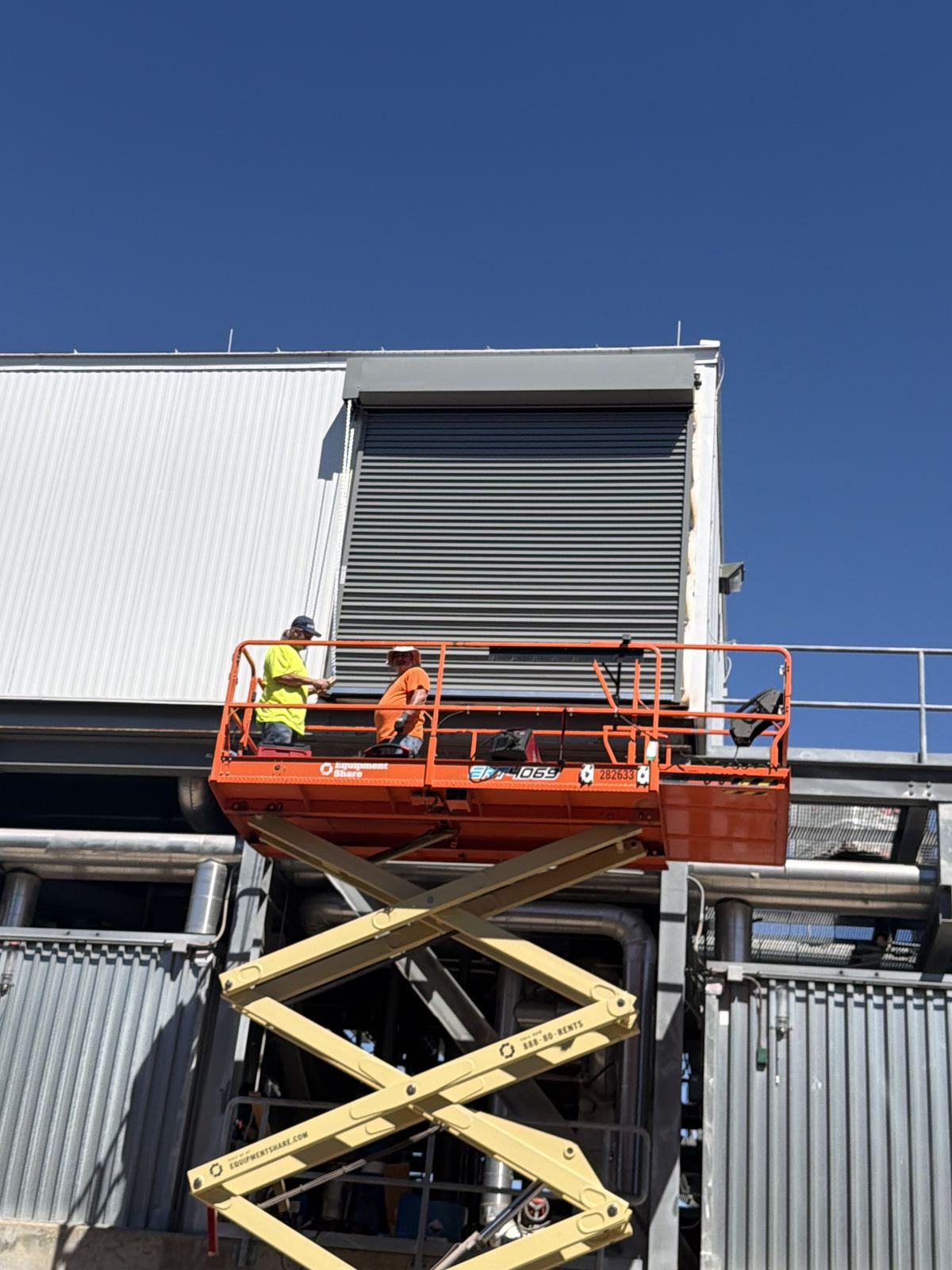 Two technicians on an orange scissor lift installing a rolling steel door high on an industrial building