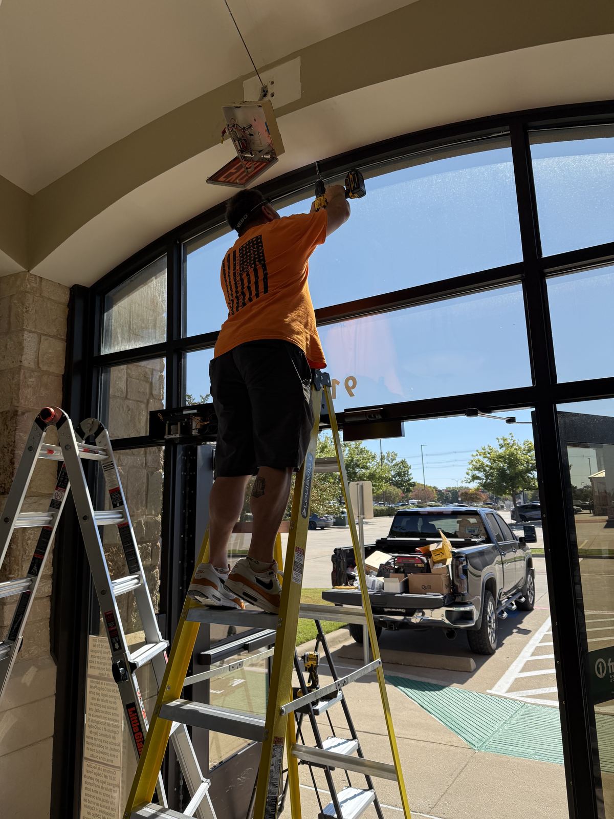 Pro Doors and Gates technician on a ladder installing an automatic door operator above an arched glass storefront entrance
